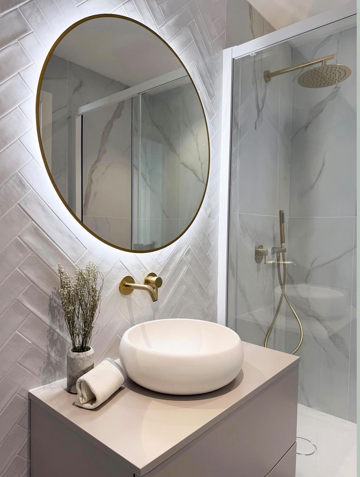 Backlit round brass mirror above a curved basin with herringbone tiling and a marble-effect shower