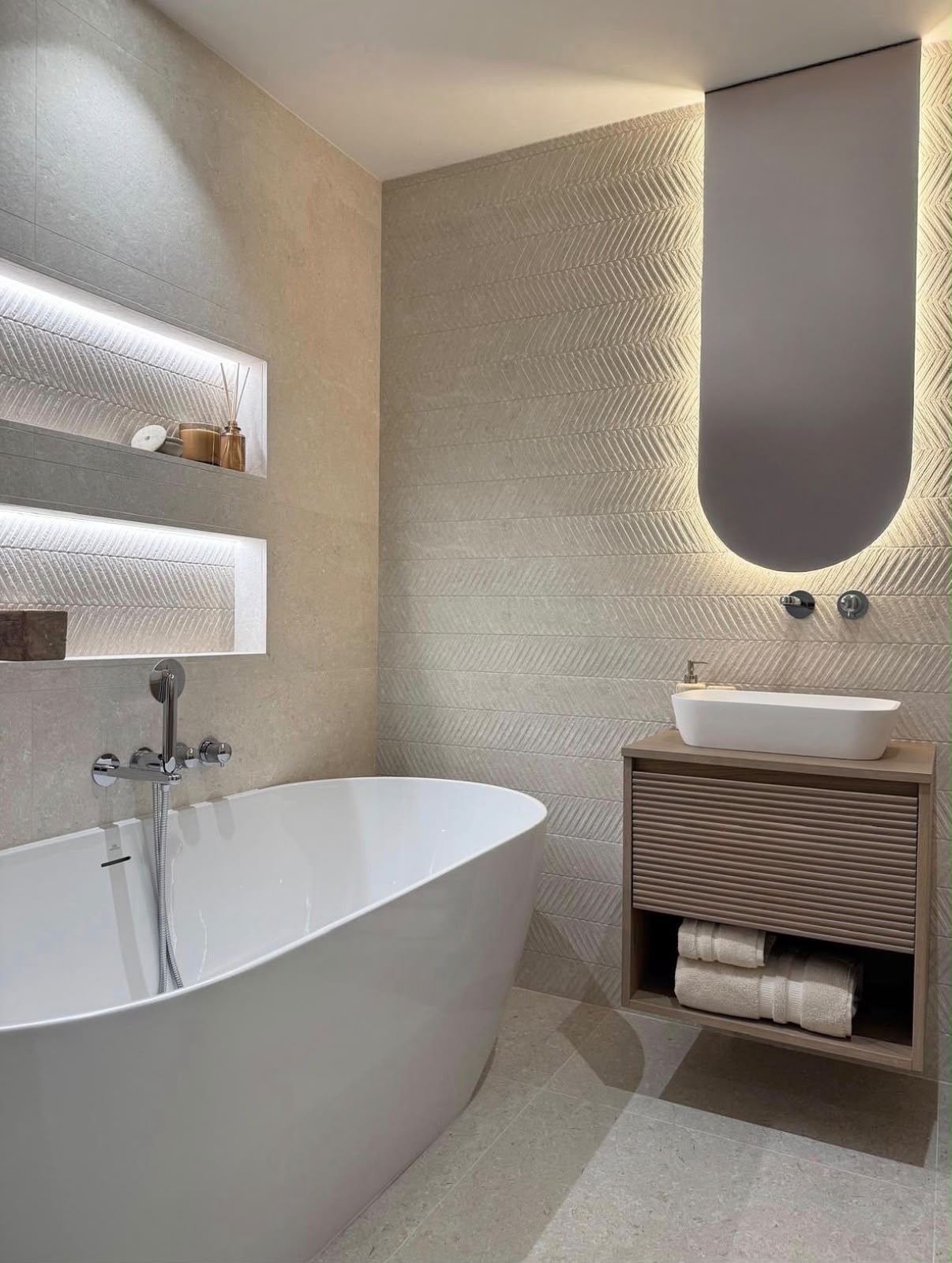 Freestanding bath with backlit niches, chevron textured stone walls and a fluted oak vanity