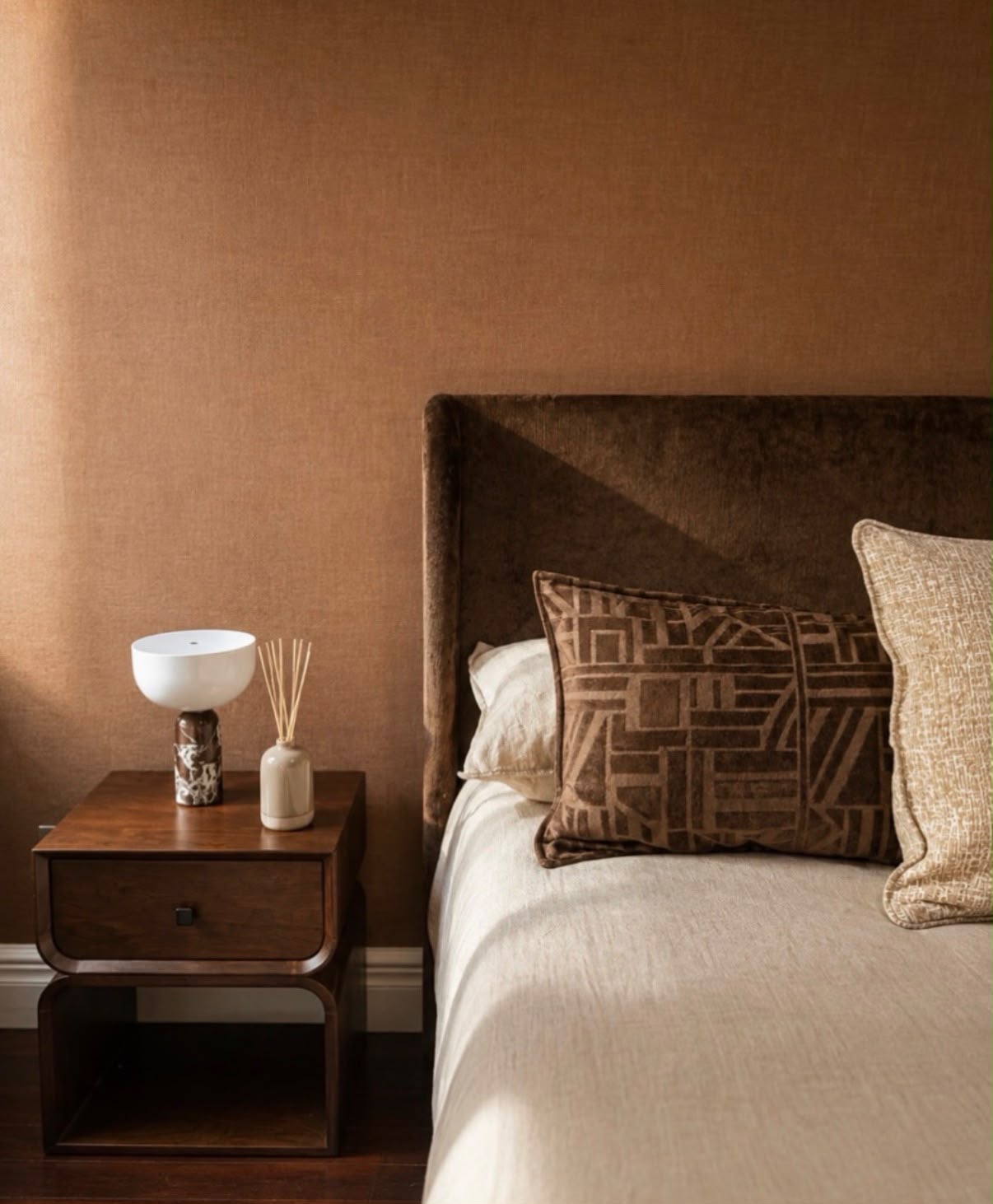 Moody bedroom detail with a chocolate velvet headboard, terracotta wall and walnut bedside table