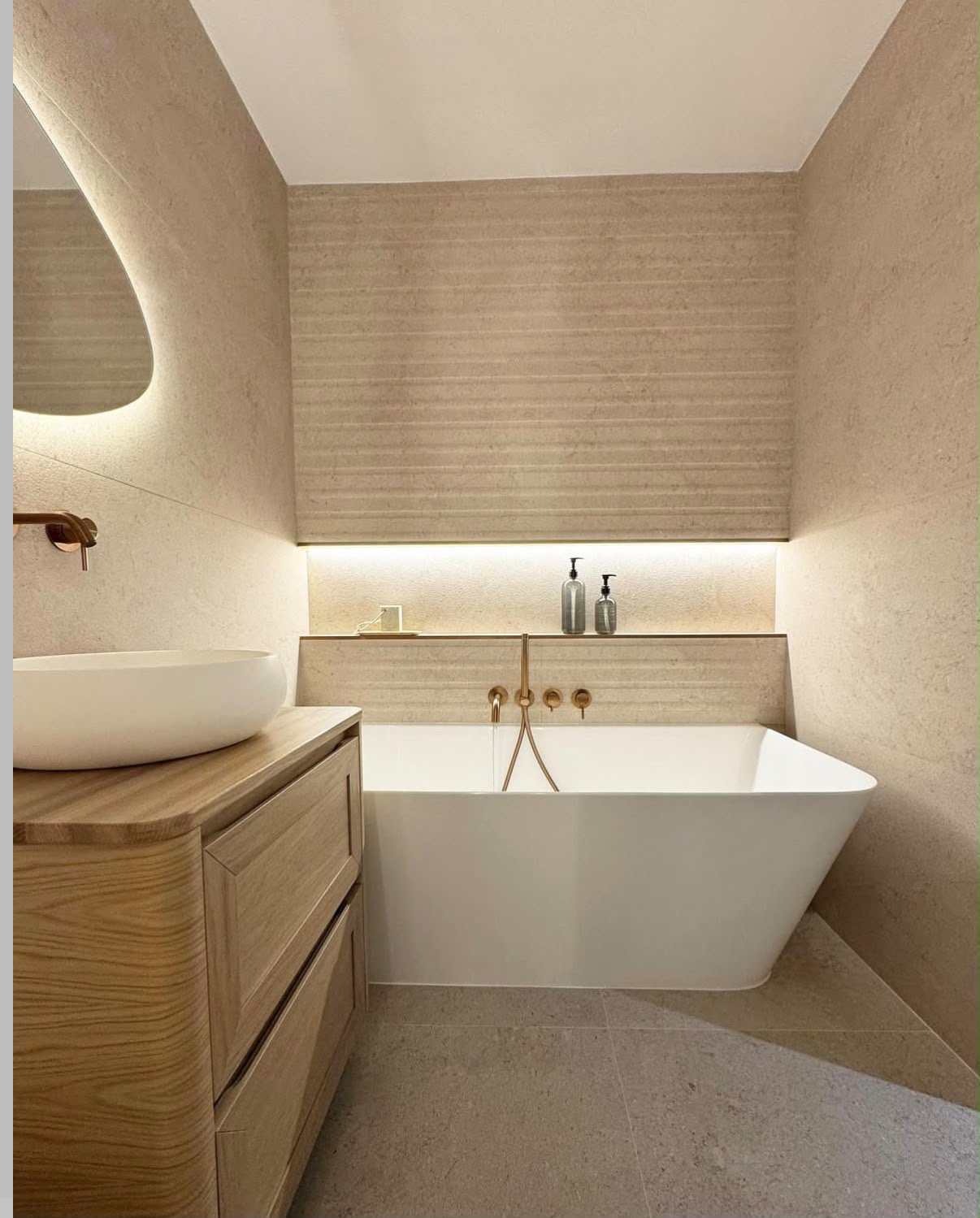 Warm travertine bathroom with a backlit organic mirror, oak vanity and brushed brass fittings